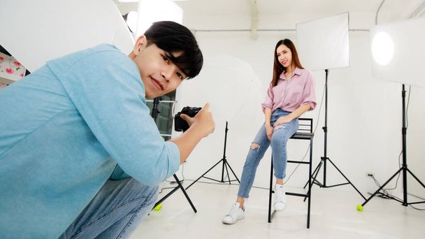 Asyalı yakışıklı erkek fotoğrafçı kadın modelin kot pantolon ve günlük gömlekle fotoğrafını çekiyor, kapalı bir stüdyoda oturuyor ve poz veriyor ışıklandırma ekipmanlarının bulanık arka planıyla. - Fotoğraf, Görsel