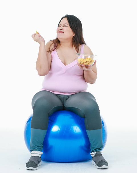 Isolated studio shot of Asian happy unhealthy overweight oversize girl in sport clothing leggings sit smiling on blue exercise fitball hold bowl of crunchy snack eating fast food on white background. - Photo, Image