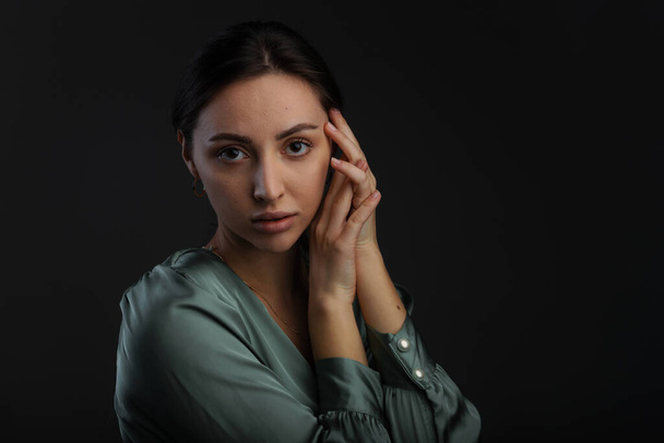 Portrait of a dark-eyed beautiful woman dressed in a green blouse, she has gold earrings and looks at camera against dark gray background, copy space - Photo, Image