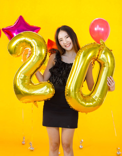 Ασιάτισσα νέα σέξι εικοσάχρονη κοπέλα με μαύρο φόρεμα που στέκεται χαμογελώντας κοίτα την κάμερα κρατώντας δύο και μηδέν χρυσά μπαλόνια glitter στα χέρια μπροστά πορτοκαλί τοίχο φόντο στο πάρτι έκπληξη. - Φωτογραφία, εικόνα