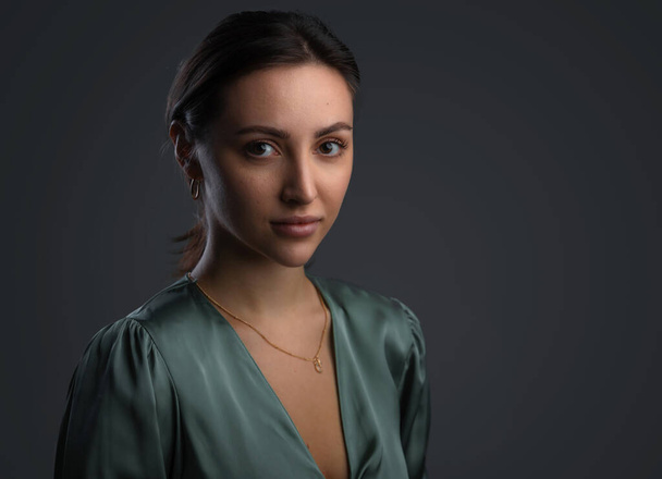 Portrait of a dark-eyed beautiful woman dressed in a green blouse, she has gold earrings and looks at camera against dark gray background, copy space - Foto, Bild