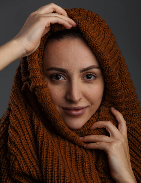 Nette junge Frau zieht sich aus und zieht sich eine Strickjacke oder einen Pullover über den Kopf, wobei ihre Arme ihr Gesicht vor dunkelgrauem Hintergrund umschließen - Foto, Bild