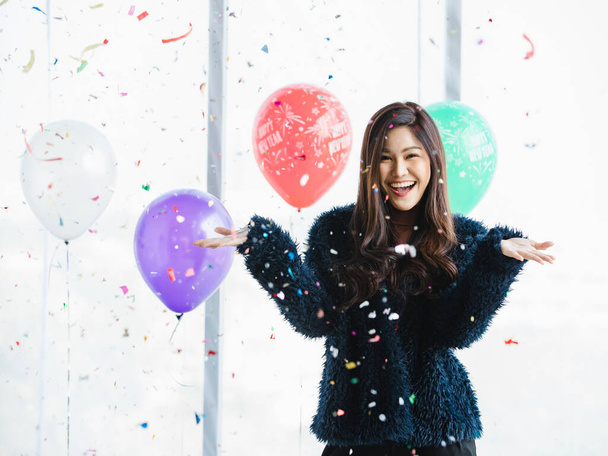 Азиатская молодая счастливая красивая женщина в синем пушистом пальто стоя улыбаясь смотреть в камеру под розовой бумаги конфетти поппер на передней красочной новогодней вечеринке празднования украшения воздушные шары фон. - Фото, изображение