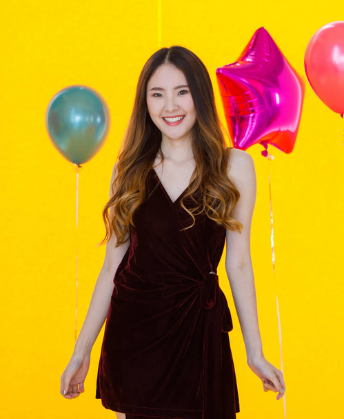 Азиатская молодая сексуальная красивая длинные волосы девушка в платье стоя улыбаясь счастливо позируя перед красочной звездой и круглой формы гелиевые воздушные шары на желтом фоне в сюрприз партии. - Фото, изображение