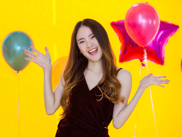 Asian young sexy beautiful long hair girl in dress standing smiling posing happily show hands up in front of colorful star and round shape helium air balloons on yellow background in surprise party. - Photo, Image