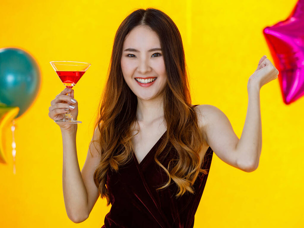 Aziatisch gelukkig mooi meisje in sexy jurk staan glimlachen kijk naar camera poseren gekruiste arm met lange cocktail glas drankje in de hand in de voorkant kleurrijke ballonnen op gele achtergrond in verrassing partij. - Foto, afbeelding