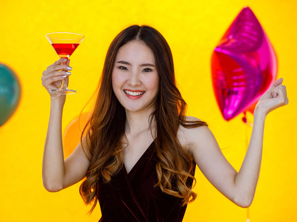 Aziatisch gelukkig mooi meisje in sexy jurk staan glimlachen kijk naar camera poseren gekruiste arm met lange cocktail glas drankje in de hand in de voorkant kleurrijke ballonnen op gele achtergrond in verrassing partij. - Foto, afbeelding