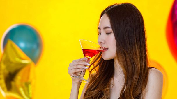 Közelkép ázsiai elég hosszú haj lány szexi ruhában áll kortyolgatva ivás piros koktél ital alkohol ital magas pohár előtt színes lufik elmosódott sárga háttér party. - Fotó, kép
