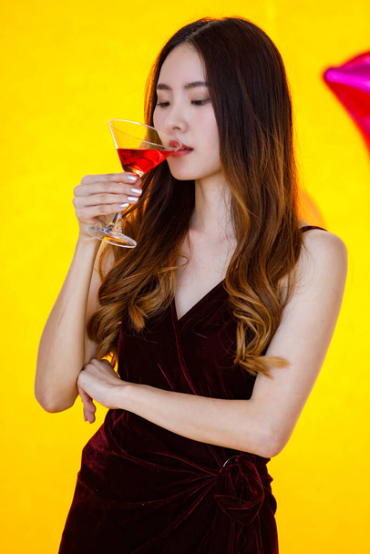 Close up shot of Asian menina cabelo muito longo em vestido sexy de pé bebendo bebida coquetel vermelho bebida alcoólica em vidro alto na frente balões coloridos no fundo amarelo desfocado na festa. - Foto, Imagem