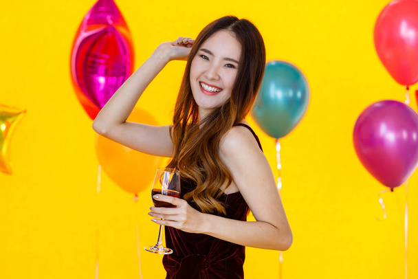 Ασιατική όμορφη κοπέλα με αυτοπεποίθηση σέξι φόρεμα στέκεται στο χέρι κρατώντας επευφημίες ψήσιμο κόκκινο κρασί ποτό αλκοόλ ποτό σε ψηλό ποτήρι μπροστά πολύχρωμα μπαλόνια σε θολή κίτρινο φόντο κόμμα. - Φωτογραφία, εικόνα