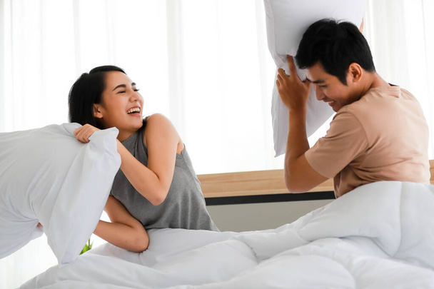 Lustige Aufnahme einer süßen lächelnden jungen erwachsenen Asiatin in lässiger Kleidung, die Spaß an einer Kissenschlacht mit ihrem Mann im Vordergrund hat. Frau und Mann spielen zusammen in der Früh - Foto, Bild