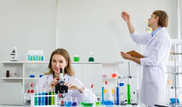 Жінка кавказька вчена дивиться через мікроскоп і вчений чоловік стоїть тримає книгу і дивиться на рідкий хімічний реагент в пробірці в лабораторії
. - Фото, зображення