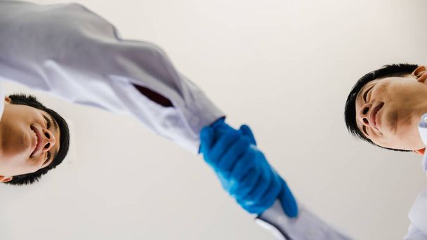From bottom view close-up focus to 2 hands are handshakes by 2 adults man who are scientist wearing white uniform and blue glove. - Foto, Imagen
