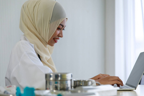 Jonge moslim vrouwelijke arts werkt met haar laptop computer op het bureau met medische apparatuur in het ziekenhuis. - Foto, afbeelding