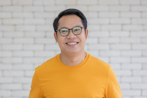 Portret dorosłego Azjaty w żółtej koszulce i okularach stojącego i uśmiechniętego patrzącego w kamerę z przyjaznym i pewnym siebie. - Zdjęcie, obraz