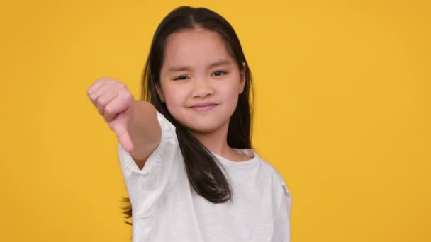 Nesnášenlivé gesto. Malá asijská dívka gestikulující palec dolů a potřásající hlavou ne, vyjadřující odmítnutí, oranžové studio pozadí - Záběry, video