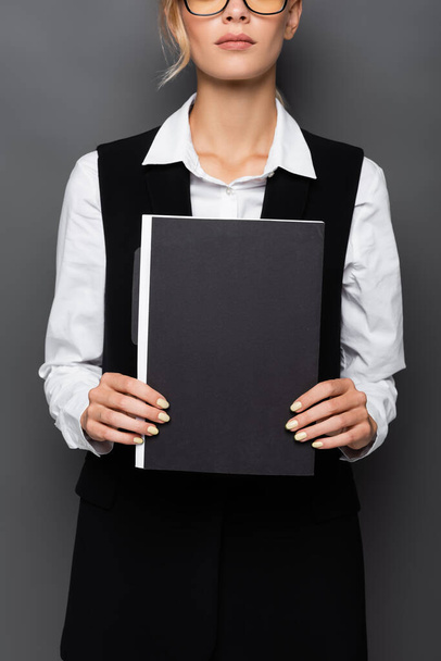 Обрезанный вид молодой предпринимательницы в очках, держащей бумажную папку, изолированную на сером  - Фото, изображение