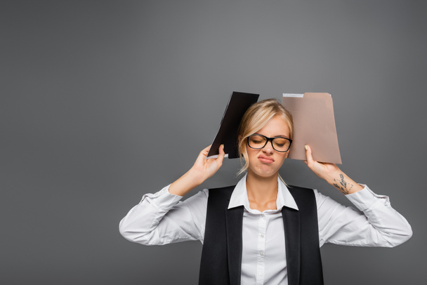 Exhausted businesswoman in eyeglasses holding paper folders isolated on grey  - Foto, afbeelding