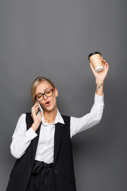 Aufgeregte Geschäftsfrau hält Pappbecher in der Hand und redet auf dem Smartphone isoliert auf grau  - Foto, Bild