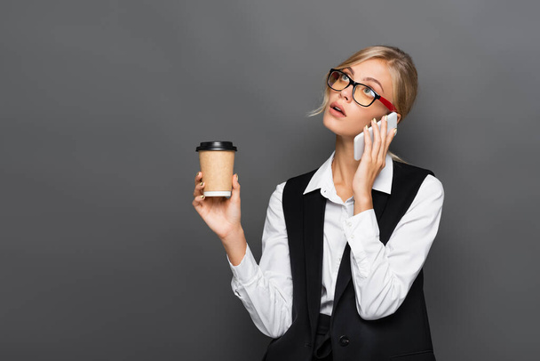 Mujer de negocios rubia pensativa hablando en el teléfono inteligente y sosteniendo café para ir aislado en gris  - Foto, imagen