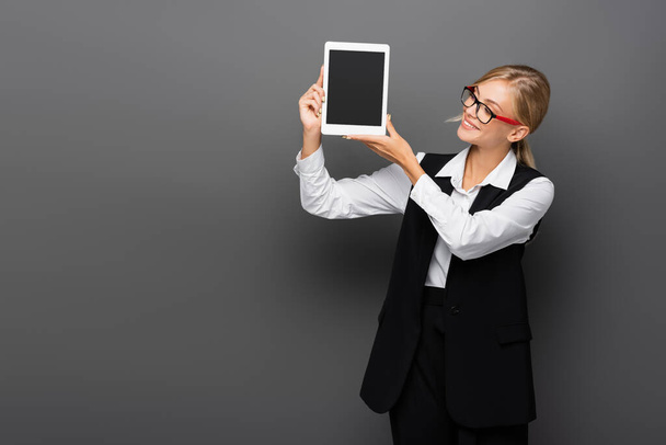 Mujer de negocios sonriente en gafas que sostiene la tableta digital con la pantalla en blanco en gris  - Foto, imagen