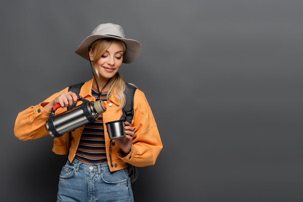 Turista sorridente con zaino contenente thermos e coppa isolati su grigio  - Foto, immagini