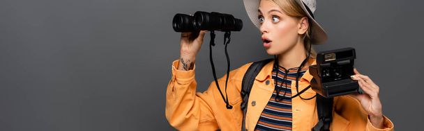 Amazed traveler holding vintage camera and looking at binoculars isolated on grey, banner  - 写真・画像
