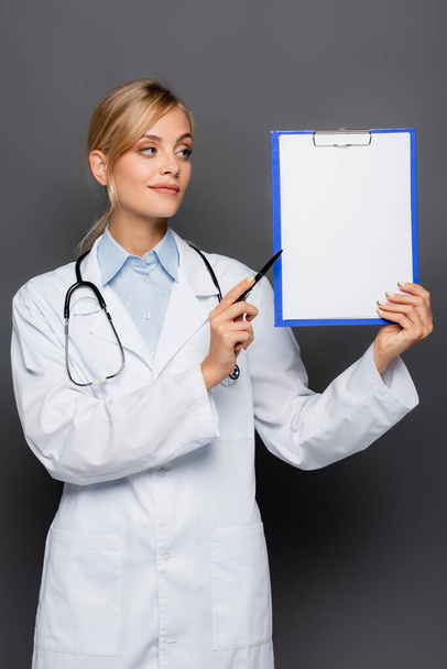 Jonge arts wijst naar klembord met kopieerruimte geïsoleerd op grijs  - Foto, afbeelding