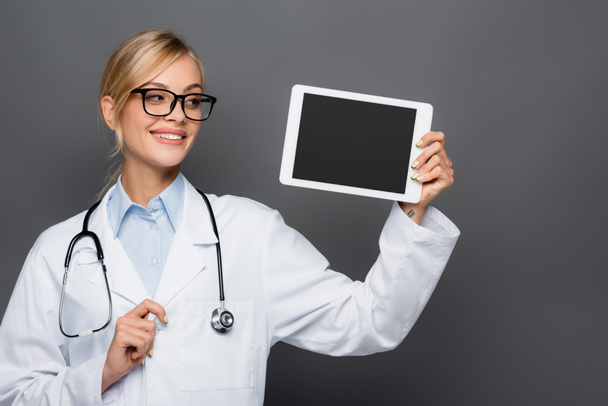 Mosolygó szőke orvos néz digitális tabletta üres képernyő elszigetelt szürke  - Fotó, kép