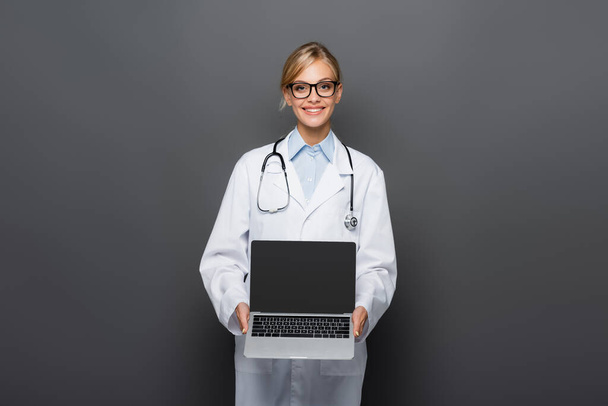 Positive doctor in white coat showing laptop with blank screen isolated on grey  - Fotoğraf, Görsel