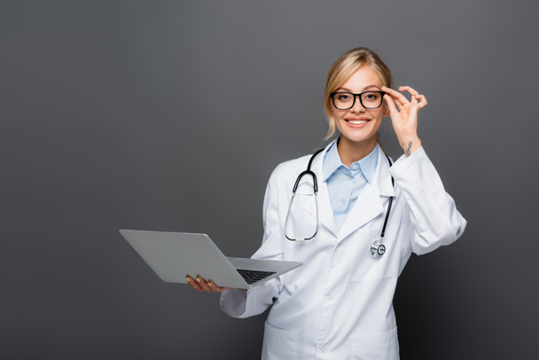 Happy doctor holding eyeglasses and laptop while looking at camera isolated on grey  - Foto, Imagem