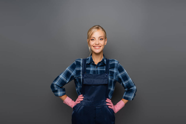 Happy workwoman in rubber gloves and overalls looking at camera on grey  - Zdjęcie, obraz