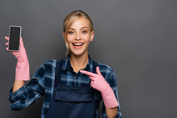 Χαρούμενη ξανθιά υδραυλικός σε λαστιχένια γάντια δείχνοντας το smartphone με λευκή οθόνη απομονώνονται σε γκρι  - Φωτογραφία, εικόνα