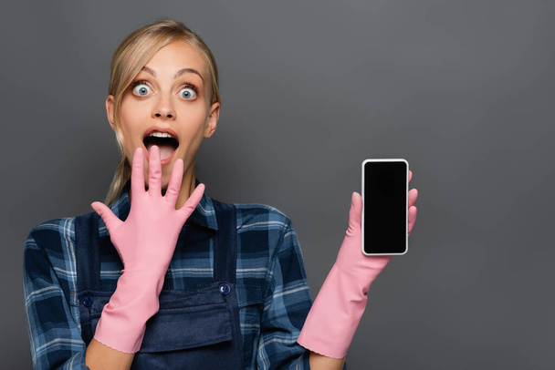 Fontanero rubio asombrado en guantes de goma sosteniendo teléfono celular con pantalla en blanco aislado en gris  - Foto, imagen