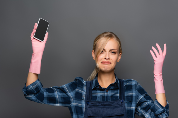 Upset plumber in overalls and rubber gloves holding smartphone with blank screen isolated on grey  - Foto, Imagem
