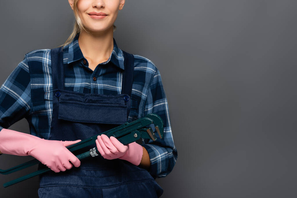 Gehakt zicht op glimlachende loodgieter in overalls en rubberen handschoenen met buissleutel geïsoleerd op grijs  - Foto, afbeelding