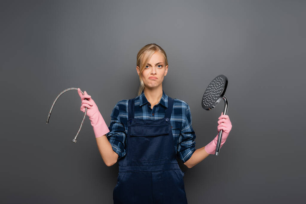 Displeased plumber in rubber gloves holding pipe and shower head on grey background - Photo, Image