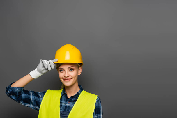 Positiver Bauarbeiter mit Helm und Warnweste blickt isoliert auf graue Kamera  - Foto, Bild