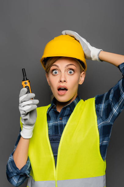 Scared builder in hardhat and safety vest holding walkie talkie and looking at camera isolated on grey  - Фото, зображення