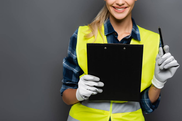 Cropped view of cheerful blonde builder in gloves and safety vest holding pen and clipboard isolated on grey  - Foto, immagini