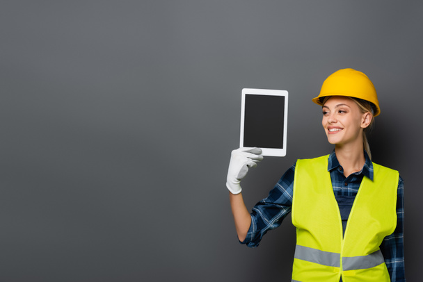 Lachende blonde bouwer in hardhat met digitale tablet met blanco scherm geïsoleerd op grijs  - Foto, afbeelding