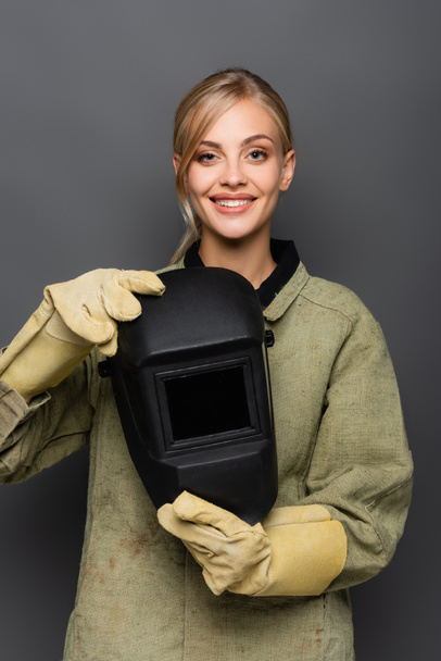 Happy blonde welder in gloves holding helmet and looking at camera isolated on grey  - Photo, Image