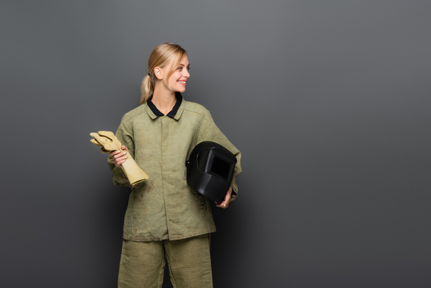Молодая блондинка сварщик держа шлем и перчатки, глядя в сторону на сером фоне - Фото, изображение