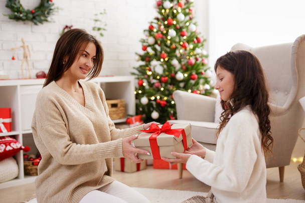 családi és karácsonyi hagyományok - fiatal gyönyörű anya karácsonyi ajándékot ad lányának a díszített nappaliban karácsonyfával - Fotó, kép