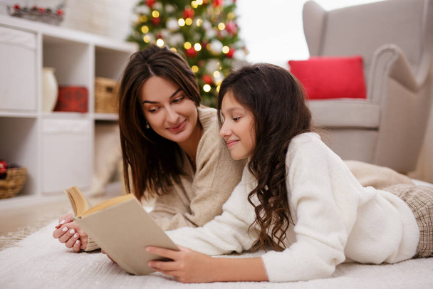 Christmas and family traditions concept - portrait of happy mother and cute daughter reading book in decorated room with Christmas tree - Photo, image