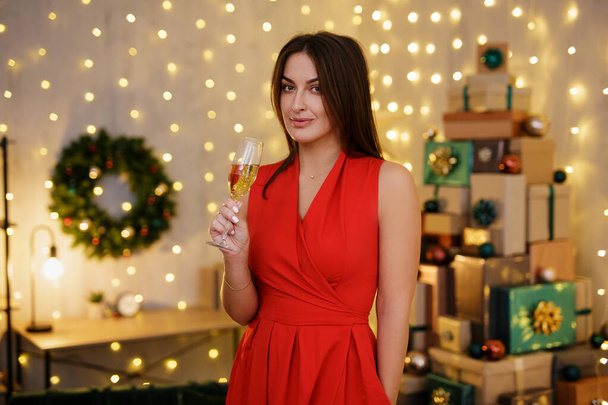 retrato de mujer hermosa en vestido rojo con copa de champán posando en habitación decorada con montón de regalos de Navidad - Foto, Imagen