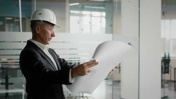 Επιτυχημένος επιχειρηματίας που εξετάζει το σχέδιο οικοδόμησης γραφείων φορώντας κράνος εσωτερικούς χώρους - Πλάνα, βίντεο