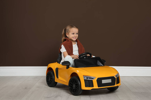 Cute little girl driving children's electric toy car near brown wall indoors - Zdjęcie, obraz