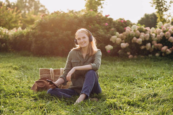 Portrait of young happy blonde woman listening to music with headphones and smiling on a green grass outdoor. Music lover enjoying music. Modern lifestyle. Online and remote education concept - Photo, Image