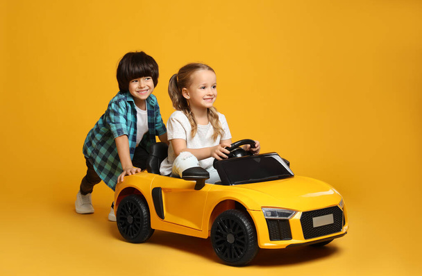 Cute boy pushing children's electric toy car with little girl on yellow background - Zdjęcie, obraz